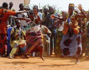 Een groep mensen voert een voodoo ritueel uit in een Afrikaans dorp. Ze dansen en zingen rond een altaar met kaarsen en offergaven, terwijl een priester de ceremonie leidt.